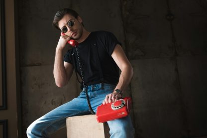handsome young man in black t-shirt and sunglasses talking by retro wired red phone